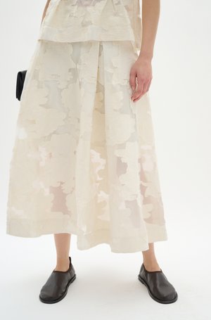 En genomskinlig, lager-på-lager maxi kjol i elfenben med texturerade blommönster, kombinerad med svarta läderskor som man glider i och en liten svart väska.