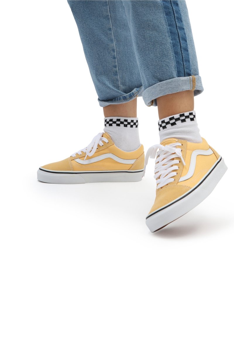 Gelbe Leinenschuhe mit weißen Akzenten und Schnürsenkeln, kombiniert mit schwarz-weiß karierten Socken. Getragen mit ausgestellten blauen Jeans.