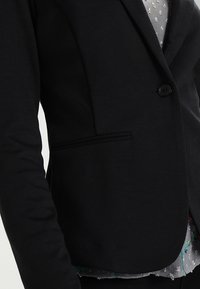 Svart skräddarsydd kavaj med en enda knapp, som har en elegant design, strukturerad axel och en sidoficka som accent. Mjuk tygkänsla.