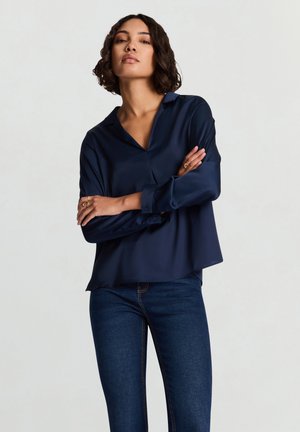 Marineblaue Satinkleidbluse mit Kragen, langen Ärmeln und Knopfmanschetten, mit einem lockeren Schnitt und glatter Textur. Kombiniert mit blauen Jeans.