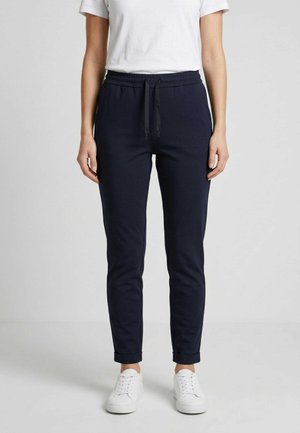 Persona che indossa pantaloni sportivi blu scuro con vita elastica e coulisse, abbinati a scarpe da ginnastica bianche e una maglietta bianca a maniche corte.