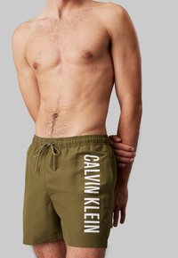 Costume da bagno verde oliva con vita regolabile tramite cordino e logo bianco "CALVIN KLEIN" verticale sulla gamba sinistra. Tessuto liscio, lunghezza fino al ginocchio.