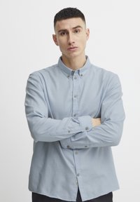 Világoskék gombos ing, gallérral, hosszú ujjakkal és barna gombokkal. Az anyag simának és enyhén texturált érzésűnek tűnik.