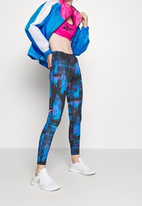 Kvinna klädd i leggings med blå, svarta och lila mönster, vita sneakers, en rosa sport-bh och en blå och vit jacka mot en vit bakgrund.