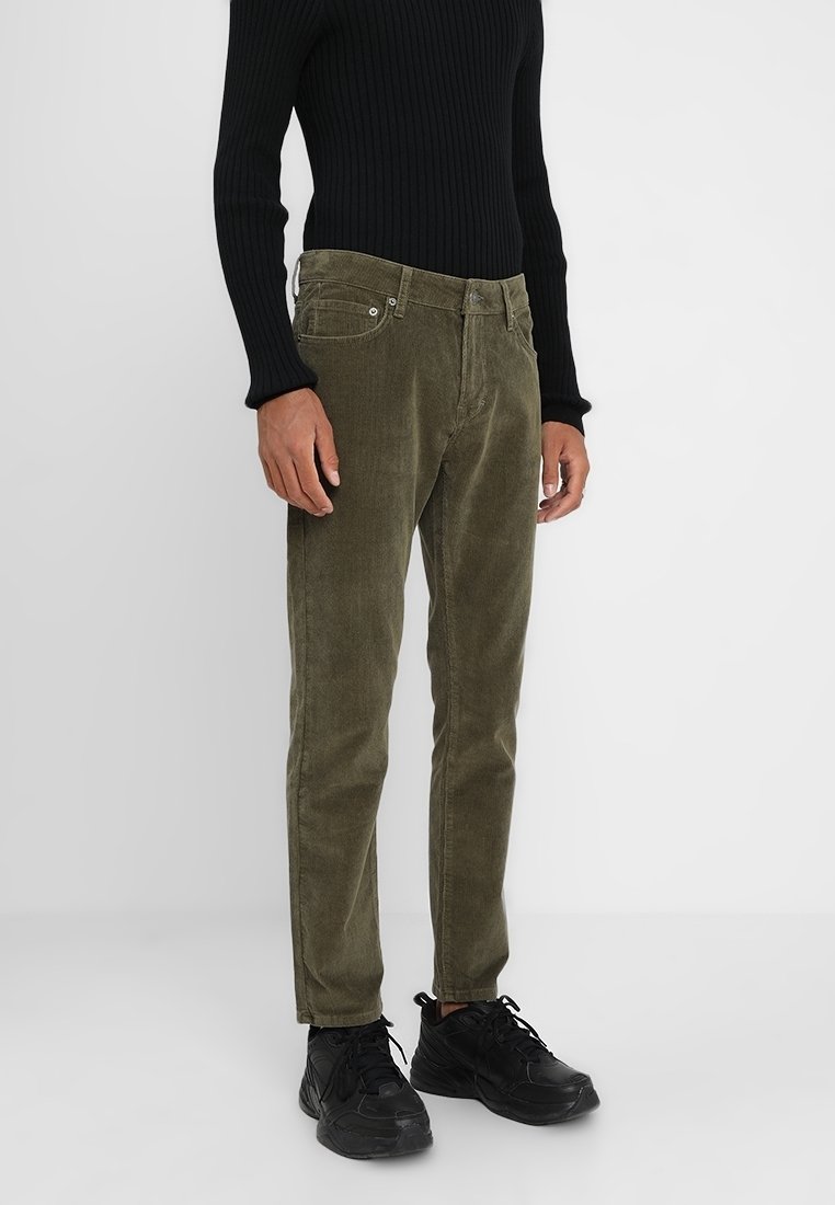 Olivově zelené kordové kalhoty se slim fit, předními kapsami a kovovými detaily, doplněné černým žebrovaným svetrem a černými botami.