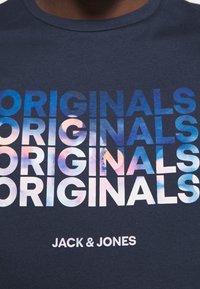 Námornícka modrá košile se slovem "ORIGINALS" opakovaným čtyřikrát v přechodu modré a růžové barvy a s nápisem "JACK & JONES" vytištěným níže.