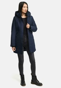 Doudoune marine avec capuche, fermeture éclair à l'avant et détails de boutons. Associée à un jean noir et des bottes noires à lacets pour un look superposé.