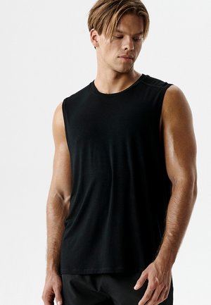 Młody mężczyzna w czarnej sportowej koszulce bez rękawów i czarnych szortach, patrzący w dół, z ramionami opuszczonymi wzdłuż ciała.