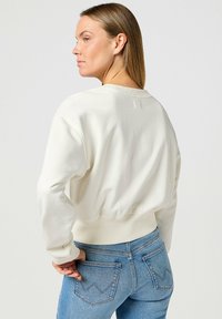 Sudadera blanca recortada con cuello redondo, dobladillo acanalado y mangas largas, combinada con jeans de mezclilla azul claro que tienen bolsillos traseros distintivos.