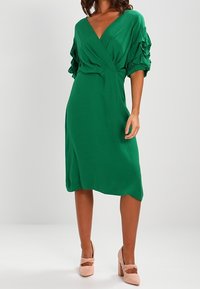 Grön omslagsklänning med puffärmar, knälång längd och mjukt tyg. Matchad med rosa klackskor med blockklack och rosettdekoration.