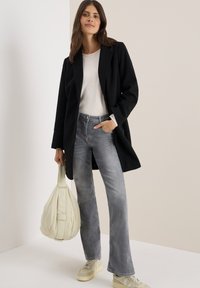 Manteau noir à la coupe classique sur un haut gris clair, associé à un jean gris délavé. Le mannequin porte un sac à bandoulière couleur crème et des baskets beiges.