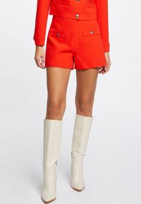 Shorts rossi su misura con tasche frontali, abbinati a stivali bianchi al ginocchio caratterizzati da un design elegante e punte affusolate.