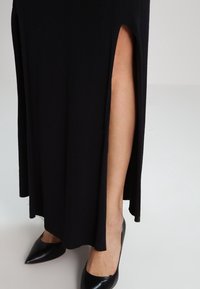 Abito lungo nero a coste con alte fessure laterali, che mette in mostra una texture liscia e una silhouette aderente. Abbinato a tacchi neri a punta.