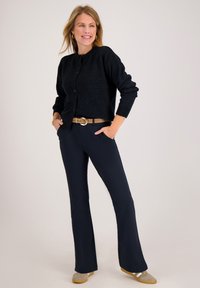 Donkergrijze cardigan met ronde hals en knoopsluiting, gecombineerd met marineblauwe wijde broek. Beige riem met gouden accenten en grijze schoenen.