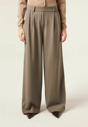 Pantalon taupe à jambes larges, taille haute et plis, porté avec un haut beige à manches longues et des chaussures pointues marron foncé.
