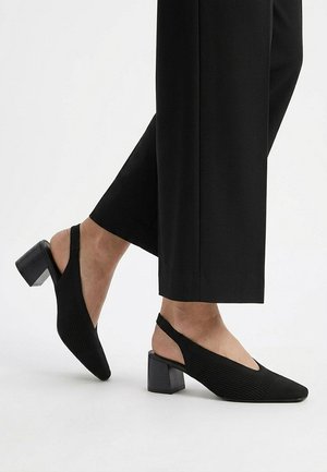 Zapatos slingback negros con tacón bajo de bloque, usados con pantalones negros recortados sobre un fondo blanco.