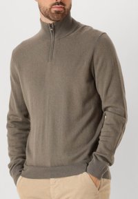 Pullover in Olivegrün, aus weichem Strickstoff. Mit einem Reißverschlusskragen und gerippten Bündchen. Kombiniert mit beige Hosen.