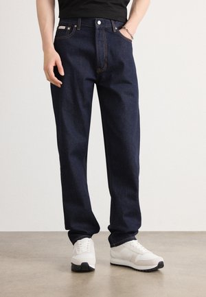 Donkerblauwe spijkerbroek met een ontspannen pasvorm, oranje stiksels, vijf zakken en een merklabel op de tailleband. Gepaard met witte sneakers.