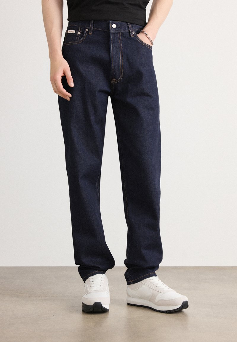 Jeans de mezclilla azul oscuro con un ajuste relajado, costuras anaranjadas, cinco bolsillos y una etiqueta de marca en la cinturilla. Combinados con zapatillas blancas.