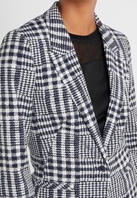 Femme portant un haut noir sous un blazer à carreaux pied-de-poule noir et blanc avec de larges revers et une fermeture à un seul bouton.