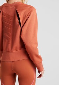 Sudadera naranja quemado con un diseño texturizado y detalle de corte en la parte trasera. Presenta puños acanalados y una forma ajustada, combinada con leggins a juego.