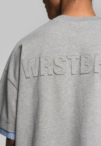 Grey oversized t-shirt with "WRSTB" stitched in a matching color; features a blue striped fabric at the cuffs for contrast.