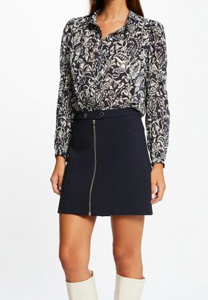 Frau trägt eine schwarz-weiße, langärmelige Bluse mit Blumenmuster, einen schwarzen Rock mit einem Reißverschluss vorne und kniehohe weiße Stiefel.