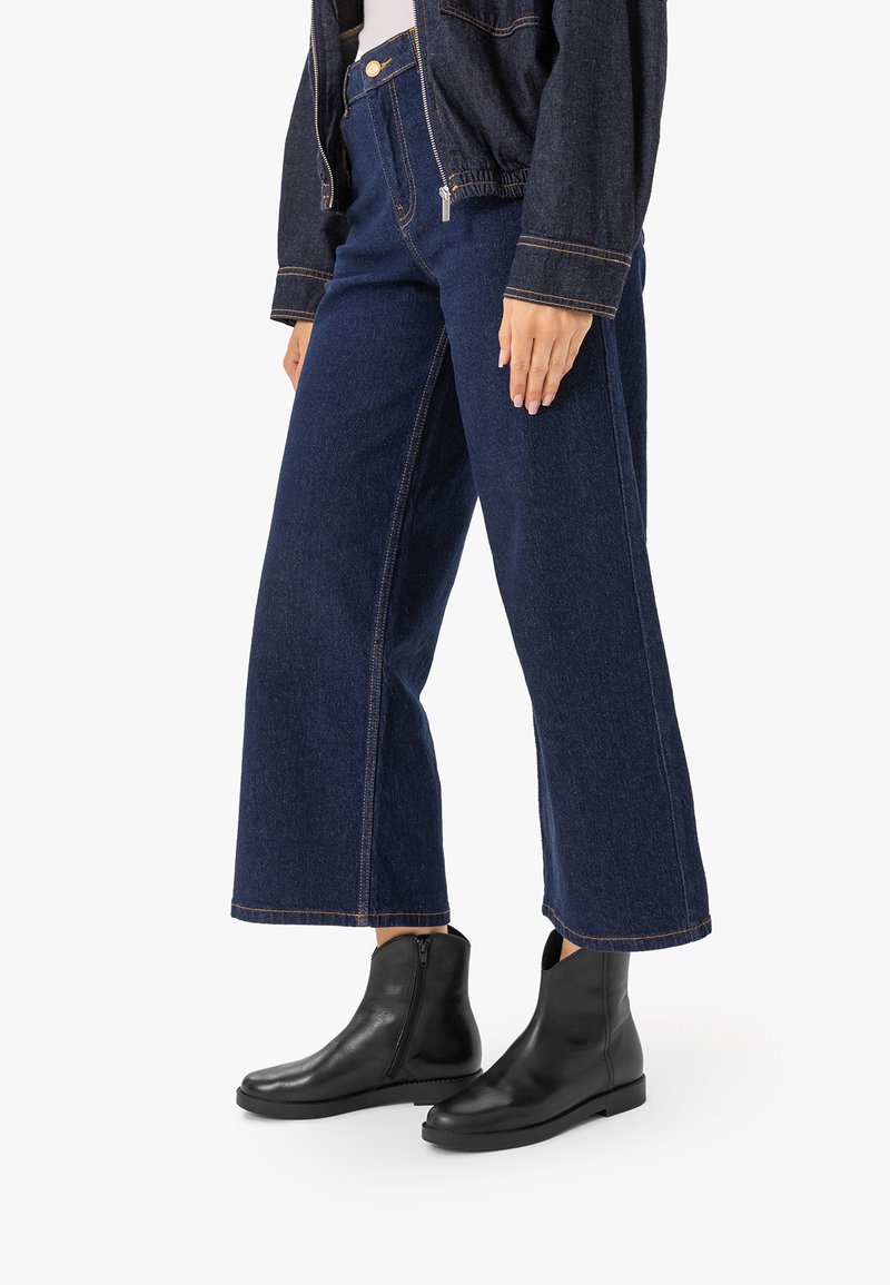 Personne portant un jean bleu marine à jambes larges, des bottines noires à fermeture éclair latérale et une veste en denim foncé sur fond blanc.