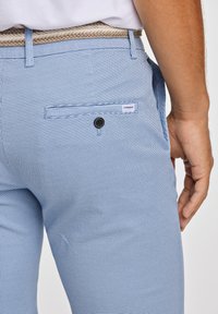 Ljust blå texturerade byxor med ett litet rutmönster, med en bakficka med en knapp och en märkesetikett. Accenterade med ett vävt beige bälte.