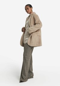 Beige Fleece-Mantel mit Kragen, großen Taschen und langen Ärmeln, getragen über einem glatten Oberteil und gemusterten weiten Hosen.