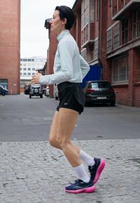 Chemise à manches longues bleu clair, shorts noirs, chaussettes blanches et chaussures de course navy avec semelles roses. Coureur dans un environnement urbain sur un pavé texturé.