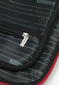 Zwarte gewatteerde tas met een gaasvak, zilveren ritssluiting en rode details. Het interieur vertoont een gestreept patroon en merknamen.