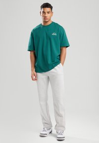 Vihreä puuvillapaita, jossa on pyöreä pääntie ja lyhyet hihat, yhdistettynä harmaisiin verkkohousuihin. Paitaan on painettu huomattava valkoinen logo rinnan kohdalle.