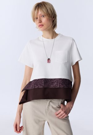 Young person with short blonde hair wears a white layered top with dark patterned hem and beige pants, posing with one hand in pocket.