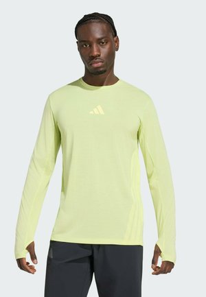 Hombre con camiseta deportiva de manga larga verde claro con huecos para los pulgares y pantalones negros, de pie frente a un fondo claro y liso.