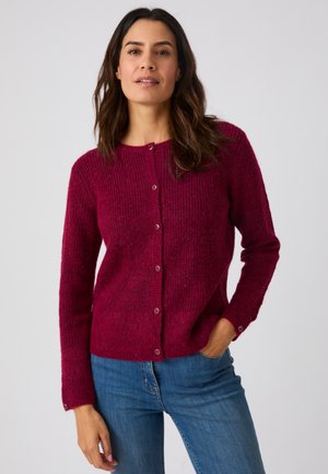 Femme aux cheveux foncés portant un cardigan en tricot bordeaux boutonné et un jean bleu, debout avec une main dans la poche sur un fond uni.