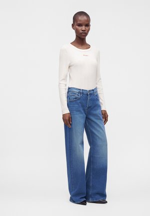 Femme portant un haut blanc à manches longues et un jean bleu taille haute à jambes larges, debout devant un fond clair uni.