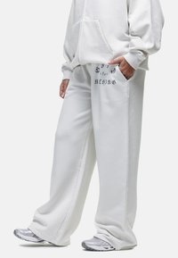 Witte oversized hoodie en bijpassende joggingbroek van zachte stof. De joggingbroek heeft pockets en bedrukte tekst nabij de tailleband. Zilveren sneakers.