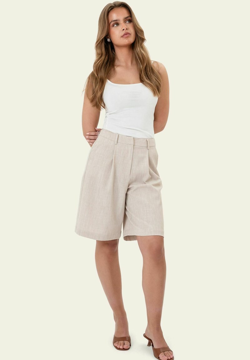 Femme portant un haut blanc sans manches, un short beige mi-genou et des talons ouverts marron, debout les mains derrière le dos.