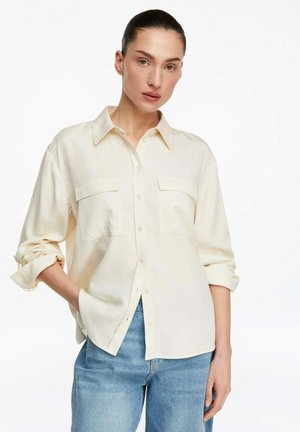 Kobieta ubrana w jasnobeżową koszulę na guziki z podwiniętymi rękawami i niebieskie jeansy, stojąca z jedną ręką w kieszeni na białym tle.