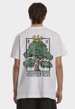 Person som har på seg en hvit skjorte med grafikk av et stort tre med en gullkrone på hodet og graffiti-tekst "Growing Root" på baksiden.