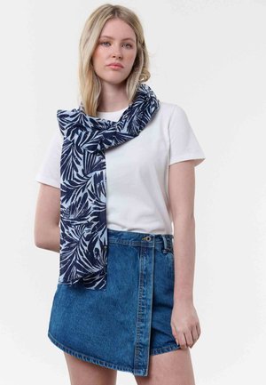 Young woman wearing white t-shirt, blue denim asymmetrical skirt, and navy blue patterned scarf draped around her neck.