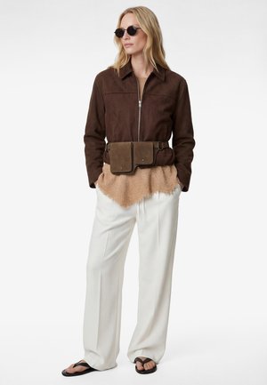 Kobieta ubrana w brązową kurtkę na suwak, beżową teksturowaną bluzkę, białe spodnie z szerokimi nogawkami, ciemną torebkę na pasku, okrągłe okulary przeciwsłoneczne i czarne sandały, stojąca z rękami w kieszeniach.