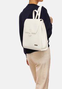 Mochila de cuero blanco con una forma estructurada, tapeta frontal, cierre decorativo y correas de hombro ajustables. Terminada con un detalle plateado.