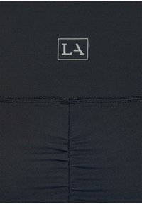 Schwarzer Stretchstoff mit geraffter Textur und einem weißen rechteckigen Logo "LA", das in der Mitte oben platziert ist. Nahtdetails am unteren Rand.