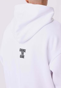 Sudadera blanca con capucha ajustable y texto negro que dice "CAMBIA EL JUEGO" en la parte de atrás. Tejido suave con un corte holgado.