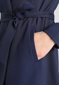 Main dans la poche avant d'un manteau bleu marine noué, à manches longues et en tissu texturé.