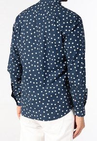 Maglietta navy con pois bianchi, colletto button-down, maniche lunghe e una texture di tessuto liscia. La vista posteriore mostra un orlo curvato.