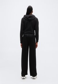 Άτομο που σ τέκεται με την πλάτη γυρισμένη, φοράει μαύρο φούτερ με κουκούλα, φαρδιά μαύρη παντελόνια και λευκά αθλητικά παπούτσια, μπροστά σε απλό ανοιχτό φόντο.