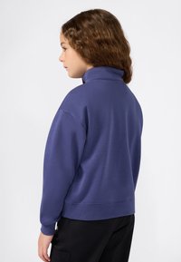 Bambino con capelli marroni ricci, che indossa un felpa blu con colletto alto e pantaloni neri, mostrato di lato e leggermente di spalle.